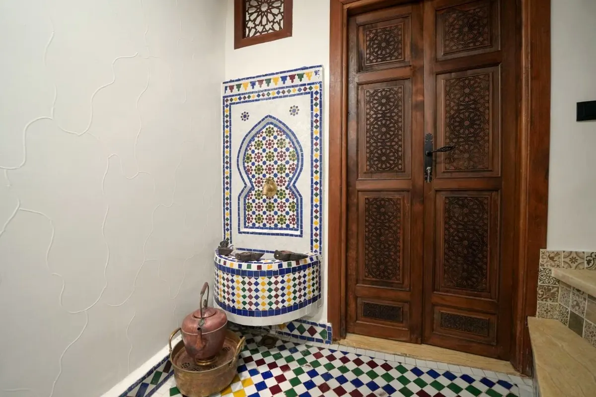 A zellige wall fountain with a brass tap, beside carved wooden doors.