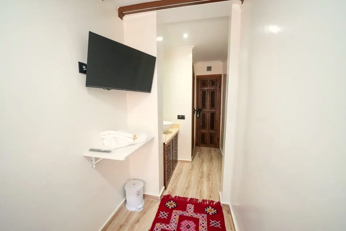 Chambre simple vue de l’entrée : télévision murale, tapis rouge et porte sculptée au fond