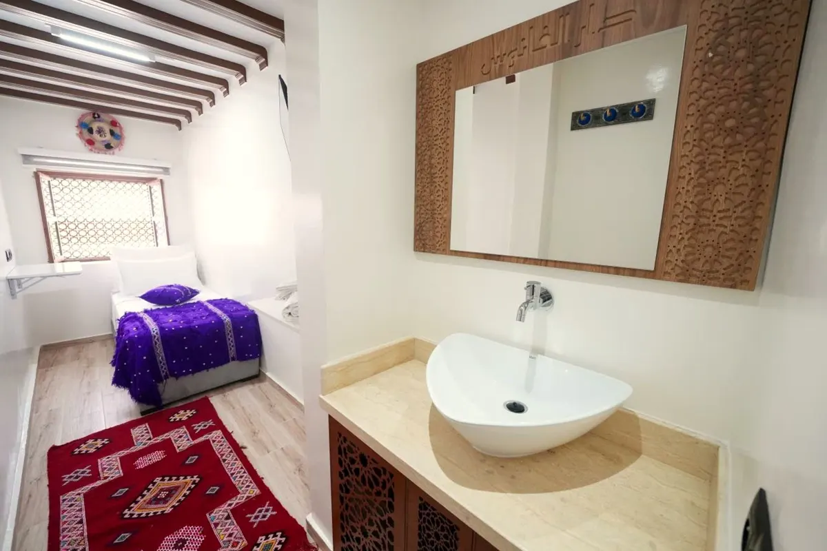 Miroir en bois sculpté et vasque posée, le lit à couverture violette au fond