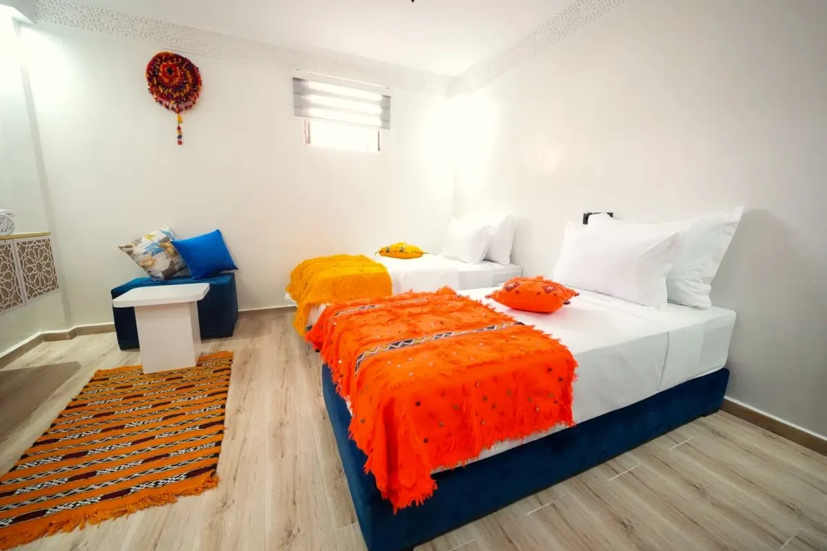 Twin room with orange blankets and a cobalt ottoman under a small high window